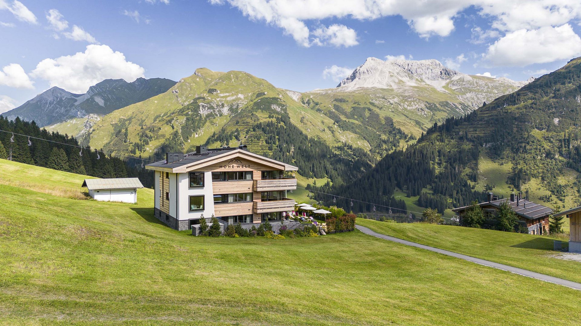 Ihr Apartment in Lech am Arlberg. Ruhe, Raum und Freiheit. Haus auf grüner Almwiese mit Bergen und blauem Himmel im Hintergrund